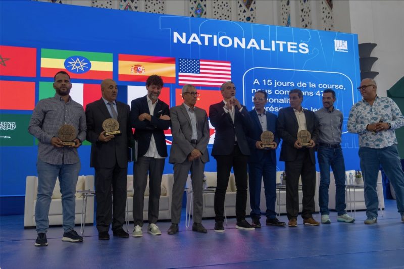 Group of men on a stage holding circular awards at an international event, with a blue backdrop displaying flags and the word NATIONALITES behind them.