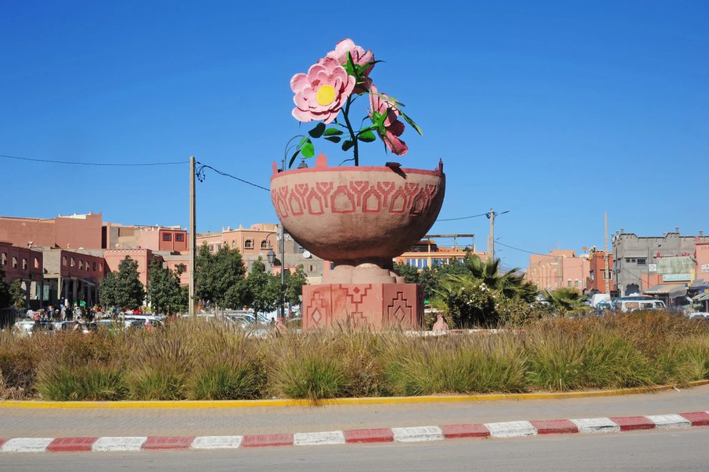 افتتاح الدورة 61 لمعرض الورد العطري بقلعة مكونة Giant pink flower sculpture rising from a red patterned planter in a town square, with pink buildings and a clear blue sky behind.
