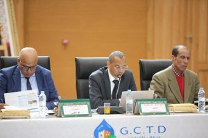Three men in suits seated at a conference table; the middle man speaks into a microphone as others listen, with nameplates and water bottles in front of them.