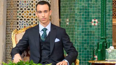 Young man in a dark suit and striped green tie sits on an ornate gold-and-cream chair in a formal setting with green tiled walls.