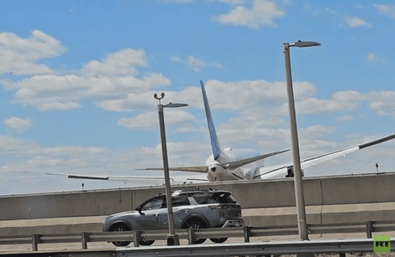 69f7d0264236044abe797e76 | MCG24 | موقع إلكتروني مغربي شامل متجدد على مدار الساعة Airliner on a taxiway behind a low barrier, with a gray SUV driving past in the foreground under a blue sky.