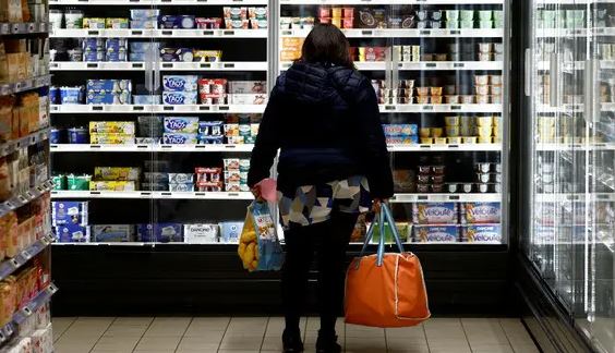 6ع7ته78ن877777777777 | MCG24 | موقع إلكتروني مغربي شامل متجدد على مدار الساعة Back view of a shopper in a supermarket dairy aisle carrying orange and yellow bags, facing refrigerated shelves labeled with dairy products.