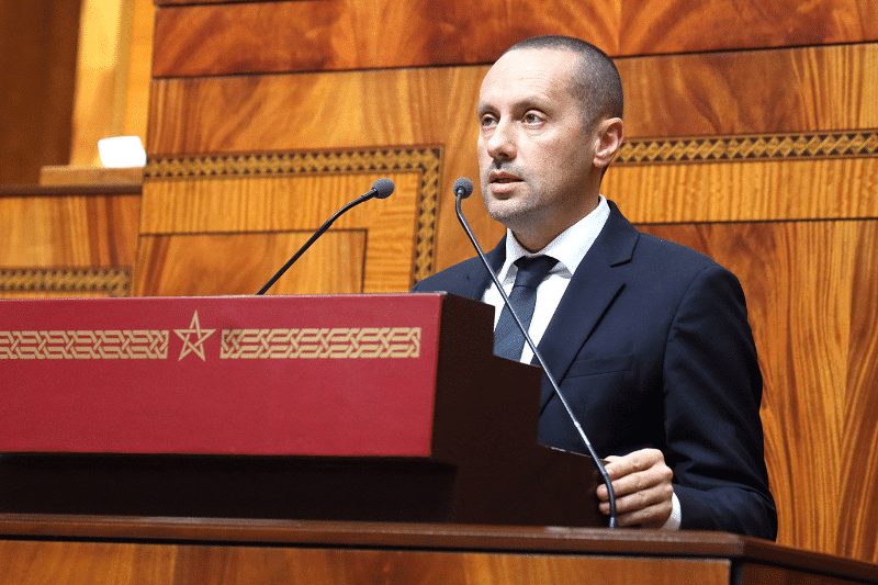 tahraoui | MCG24 | موقع إلكتروني مغربي شامل متجدد على مدار الساعة Man in a dark suit delivering a formal speech at a wooden podium with two microphones in a large wood-paneled chamber; calm, focused expression while addressing an audience.