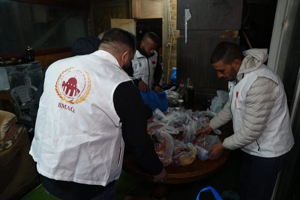 DSC00707-scaled | MCG24 | موقع إلكتروني مغربي شامل متجدد على مدار الساعة Volunteer group wearing white vests with BMAQ logo sorting donated bags of supplies on a table indoors.