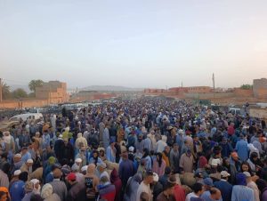 FB_IMG_1779352296792 | MCG24 | موقع إلكتروني مغربي شامل متجدد على مدار الساعة Large crowd of people gathered in a dusty desert town street, many wearing traditional robes and head coverings, suggesting a market or public event at dusk.