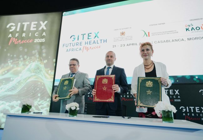 Three professionals on a stage hold red, green, and blue folders during a conference, with GITEX Africa Morocco banners in the background.