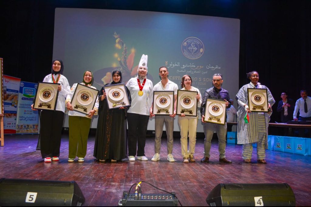 من التكوين إلى الإبداع.. جمعية إسراء تصنع جيلا جديدا من نجوم الطهي Group of people on a stage posing with large framed awards, some in chef coats and uniforms, backdrop with logos.