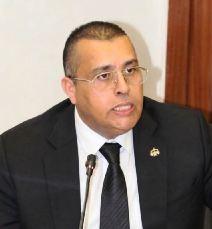 IMG-20260515-WA00711 | MCG24 | موقع إلكتروني مغربي شامل متجدد على مدار الساعة Man in a dark suit and tie speaking into a microphone at a formal meeting, wearing glasses and a lapel pin.