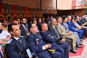 Group of military and civilian officials seated in a row at a formal ceremony under a decorated tent.