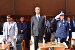 Dignitaries stand at a formal ceremony: man in a black suit at center, others in suits, a uniformed officer saluting on the right.