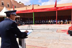 Uniformed officer gives a speech at a podium during a formal outdoor ceremony, with officials seated under a red tent in the background.