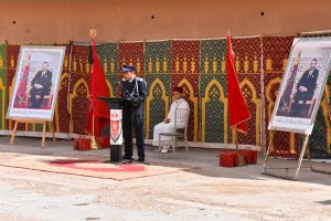 A uniformed officer speaks at a podium during a formal outdoor ceremony, with a child in white seated beside him and large portraits on easels in the background.