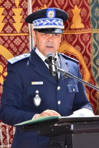 Police officer in a navy uniform and ceremonial hat speaks at a podium with a microphone; name badge shows A. Azhar, ornate backdrop behind him.