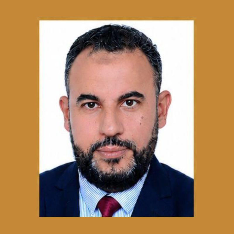 Professional headshot of a bearded man in a navy suit and red tie, with a white background and cropped framing.