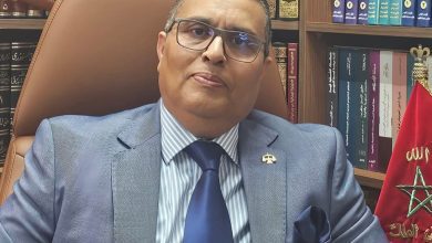 Professional man in a blue suit and striped shirt sits in a leather chair in a book-filled office.