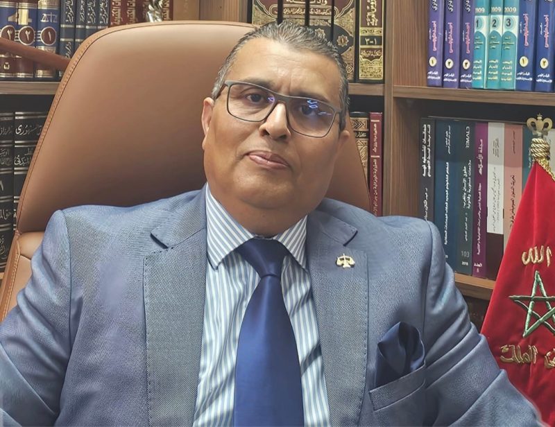 IMG-20260524-WA0037 | MCG24 | موقع إلكتروني مغربي شامل متجدد على مدار الساعة Professional man in a blue suit and striped shirt sits in a leather chair in a book-filled office.