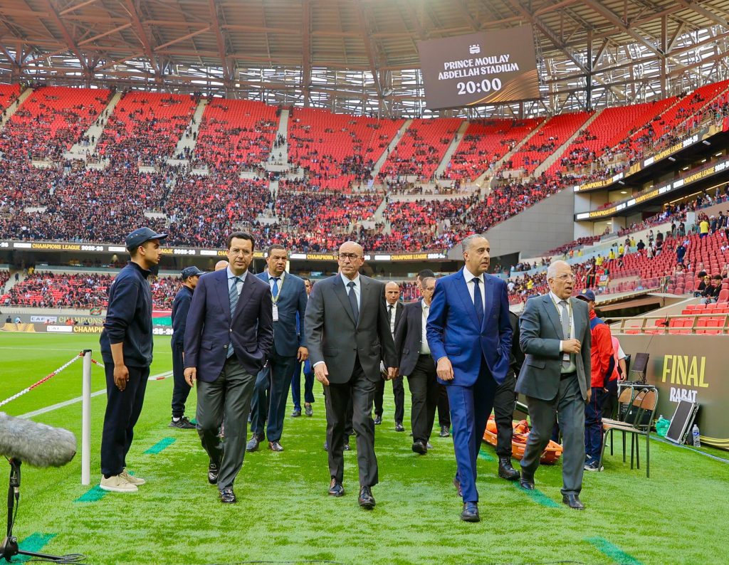 Group of men in suits walking along a soccer field in a large stadium with red seats and a digital scoreboard overhead; crowd seated in the stands behind them.