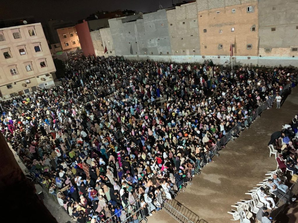 أيت ملول يحتضن النسخة الأولى من مهرجان بوجلود Aerial night-time view of a large crowd gathered in an open area behind metal barriers, with surrounding apartment buildings and dim street lighting.