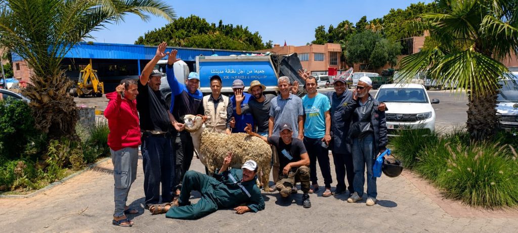 IMG-20260531-WA0193 | MCG24 | موقع إلكتروني مغربي شامل متجدد على مدار الساعة Group of men posing with a sheep in a sunny outdoor lot, with trucks and palm trees in the background.