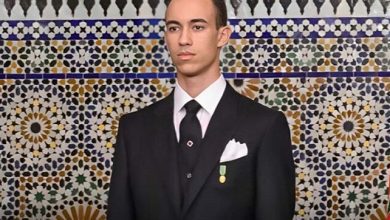 Young man in a black suit with a white pocket square and medal, standing before a colorful mosaic wall.