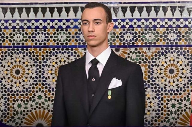 Young man in a black suit with a white pocket square and medal, standing before a colorful mosaic wall.