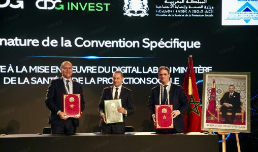 MAP9876546_png | MCG24 | موقع إلكتروني مغربي شامل متجدد على مدار الساعة Three men in suits hold red and green folders on stage during a formal signing ceremony, Moroccan flag visible nearby.