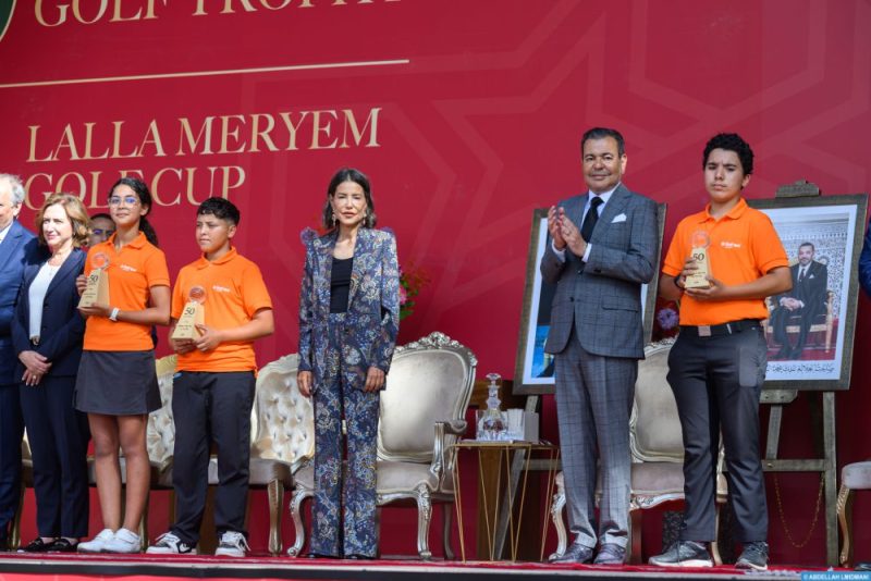 Young award recipients in orange shirts hold trophies on stage beside suited officials against a red backdrop of the ceremony.