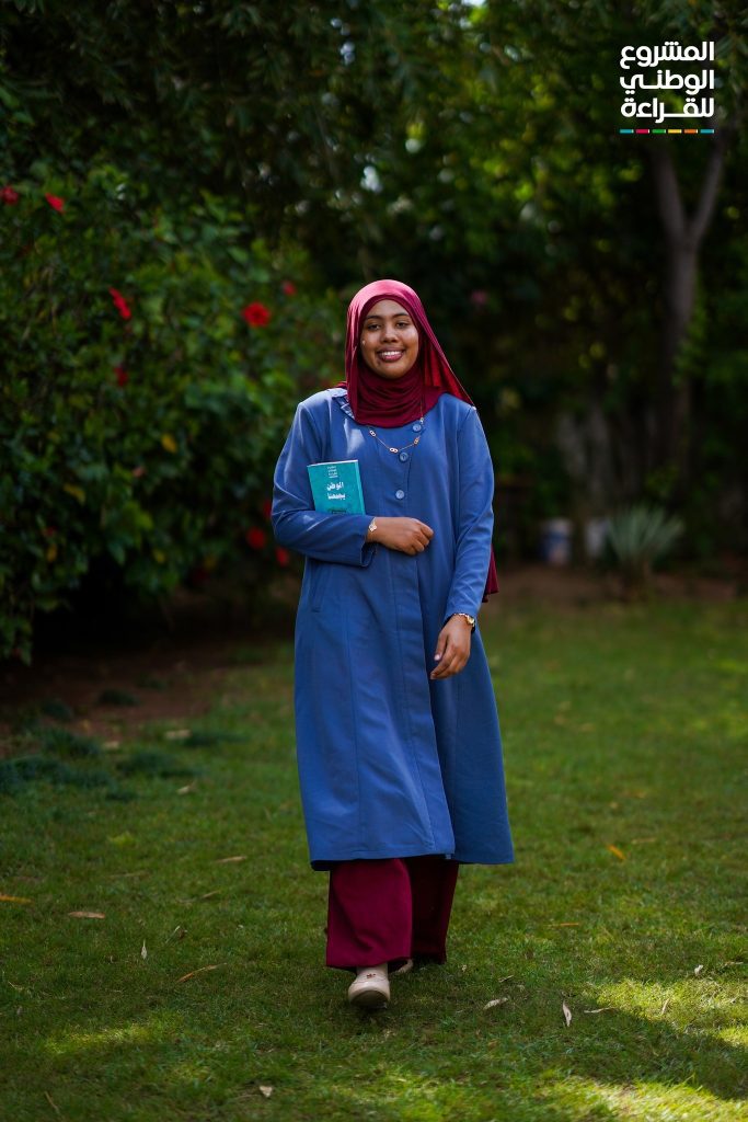Smiling woman in a red hijab and blue coat stands on grass outdoors, holding a teal booklet.