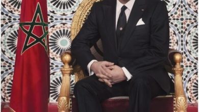Man in a dark suit sits on an ornate gold chair beside the Moroccan flag, with a decorative tiled backdrop and Arabic calligraphy.