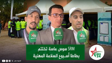 Three men speaking to reporters at an outdoor event; green MCG24 microphones in front, tents and a crowd in the background.