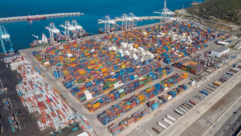 Tanger-Med | MCG24 | موقع إلكتروني مغربي شامل متجدد على مدار الساعة Aerial view of a busy container terminal with colorful stacked shipping containers, gantry cranes, and the blue sea nearby