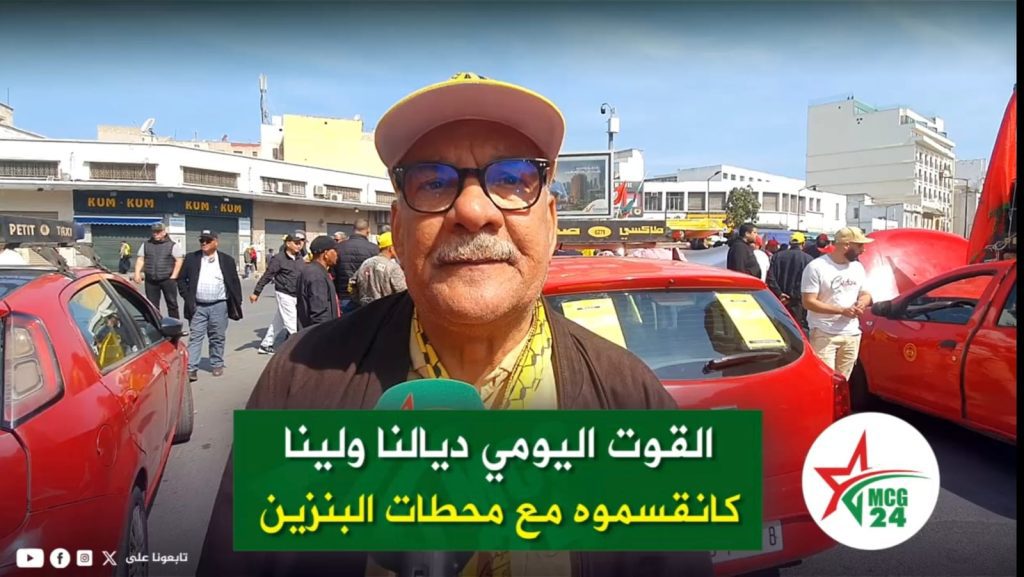 Close-up of a man with glasses and a pink cap speaking into a teal microphone at a street car event, with a green Arabic caption banner in the background and red cars nearby.