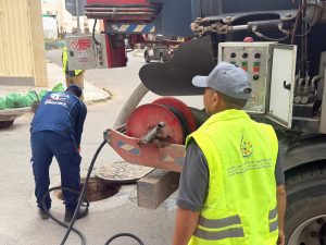 Two workers in high-visibility vests inspect a maintenance truck and hoses near an open manhole on a street, performing sewer work.