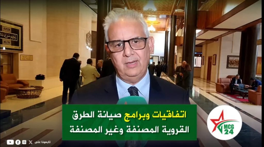 البرنامج المندمج للتنمية المحلية يفتح الباب على جيل جديد من الطرق Man in a suit and glasses giving a televised interview in a hotel lobby, with a green Arabic news caption at the bottom and a logo on the right.