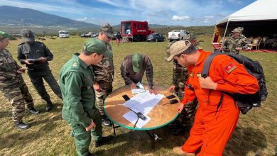 Soldiers in green and camouflage uniforms gather around a round table on a grassy field, reviewing maps and papers.