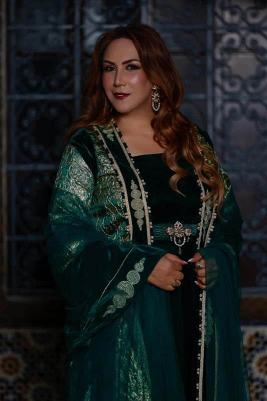 Elegant woman in a dark emerald green, embroidered gown with silver trim and statement earrings, standing with hands clasped.