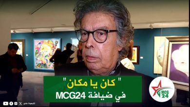 Close-up of an older man with gray hair and black glasses in an art gallery, colorful paintings in the background, and a green caption bar with Arabic text across the chest area.