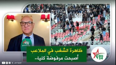 News thumbnail: man in a suit with a green mic (MCG24) speaking, stadium stands in background, green banner with Arabic text about rejecting stadium hooliganism entirely.