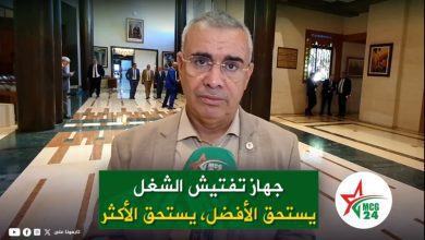 Man in a beige suit being interviewed in a marble lobby, with a green caption banner in Arabic and the MCG24 logo visible on the right.
