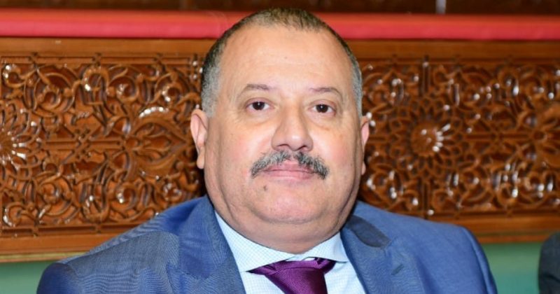 WhatsApp Image 2026-05-14 at 090203 | MCG24 | موقع إلكتروني مغربي شامل متجدد على مدار الساعة Middle-aged man with a mustache in a blue suit and burgundy tie, seated before a carved wooden backdrop.
