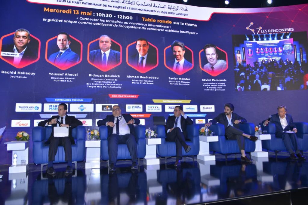 Panel of five men in suits seated on a blue stage during a formal conference, with a large backdrop of portraits and event text behind them.