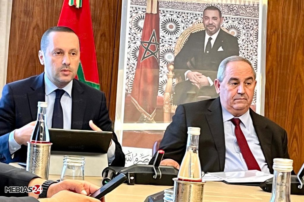 WhatsApp Image 2026-05-15 at 134456 | MCG24 | موقع إلكتروني مغربي شامل متجدد على مدار الساعة Two men in suits at a formal meeting, with a Moroccan flag and a large portrait behind them.