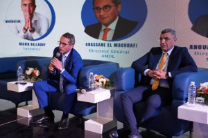 WhatsApp Image 2026-05-15 at 223713 | MCG24 | موقع إلكتروني مغربي شامل متجدد على مدار الساعة Two businessmen in blue suits sit on a panel stage, speaking into a microphone, with floral centerpieces on small tables nearby.
