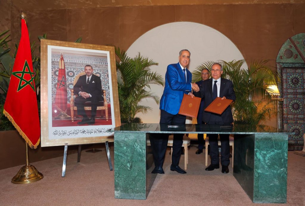 WhatsApp Image 2026-05-19 at 200649 | MCG24 | موقع إلكتروني مغربي شامل متجدد على مدار الساعة Two men in suits shake hands while holding signed documents during a formal ceremony, with the Moroccan flag and a large framed portrait nearby.