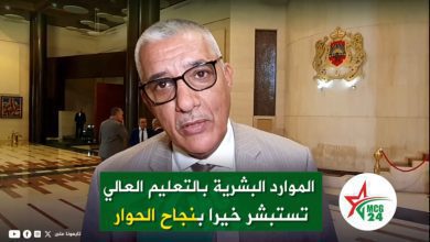 Man in a light gray suit and black glasses speaks in a lobby with a green Arabic caption overlay about higher education resources and dialogue, and a logo reading MC G24.