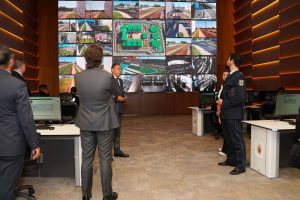 Control room with a wall of screens showing urban surveillance and road networks as staff discuss at desks.