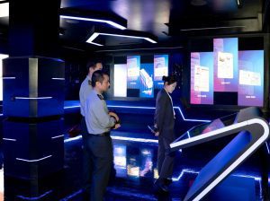 Two uniformed men and a woman in a suit study a futuristic blue-lit exhibition with a touchscreen podium.