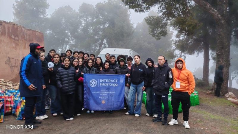 Group of students posing outdoors with a blue INTERACT banner in a misty, wooded area; a van behind them and backpacks on the ground.