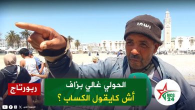 Outdoor news interview on a sunny beach; man points toward the camera with a green Arabic caption banner and reporter mic in view.