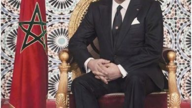 Portrait of a man in a dark suit seated on an ornate gold chair beside a Moroccan flag, with Arabic script below.